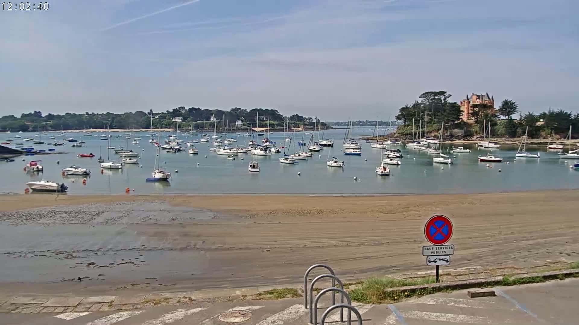 A calm, sunny day shows a sandy beach and a harbor filled with numerous sailboats and motorboats, with a large building visible on a small, tree-covered peninsula in the background.