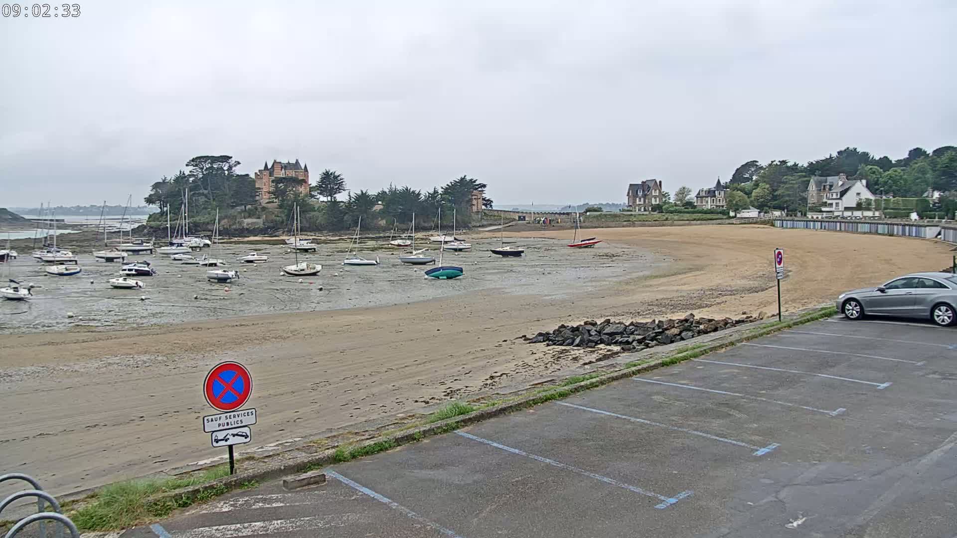 A sandy beach with numerous sailboats resting on the exposed seabed, a chateau-like building on a small island in the background, and a car parked in an empty lot under an overcast sky.