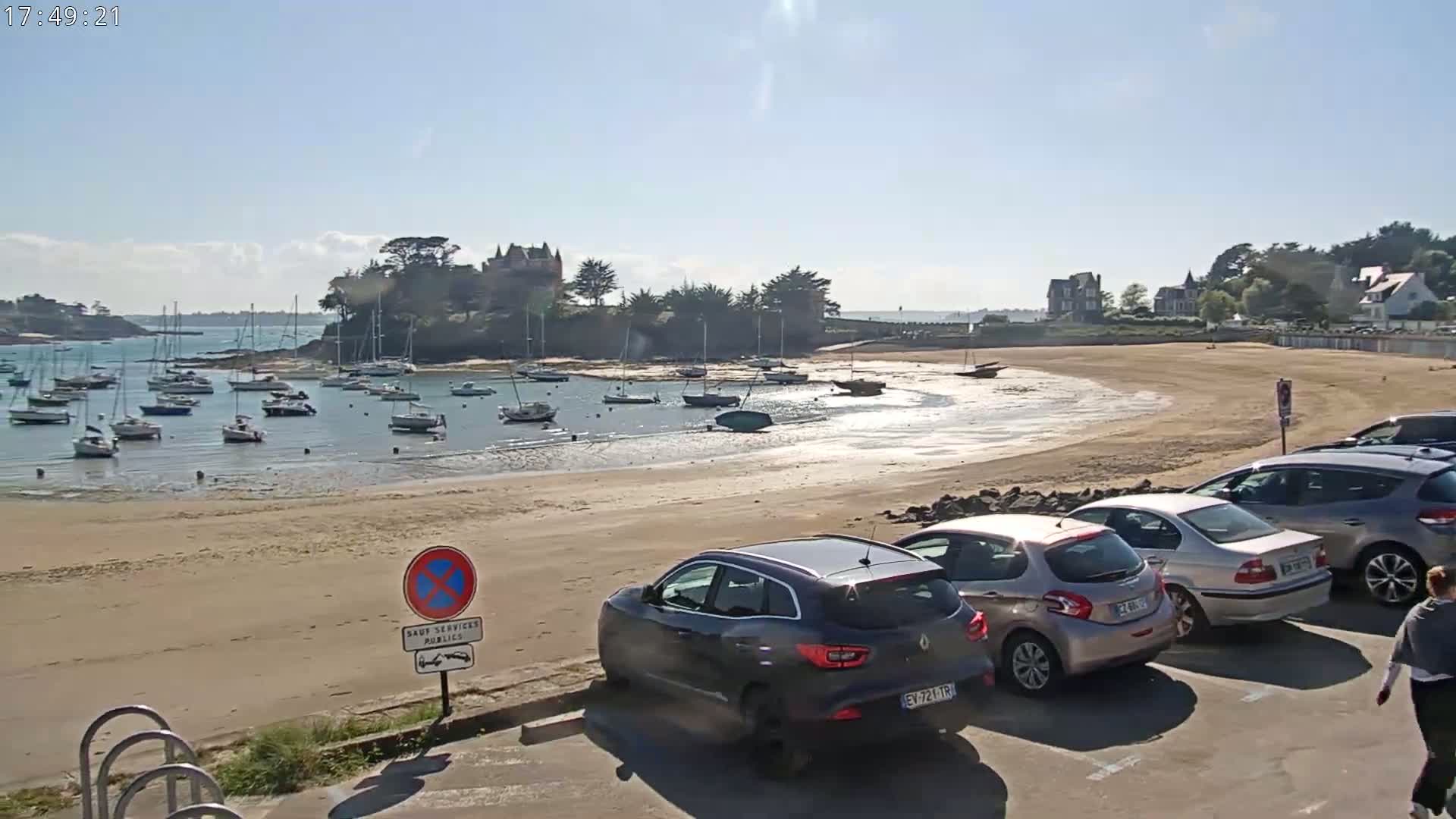 A sandy beach with several sailboats in the calm water, a few cars parked nearby, and a building on a small island in the background under a sunny sky.