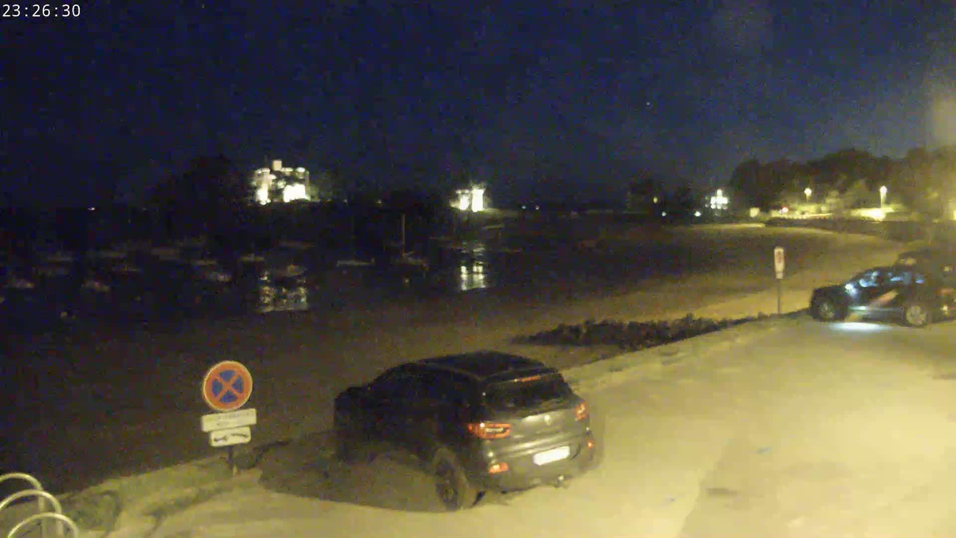 At night, two cars are parked in a lot near a dark, calm body of water, with faintly lit buildings and trees in the background.