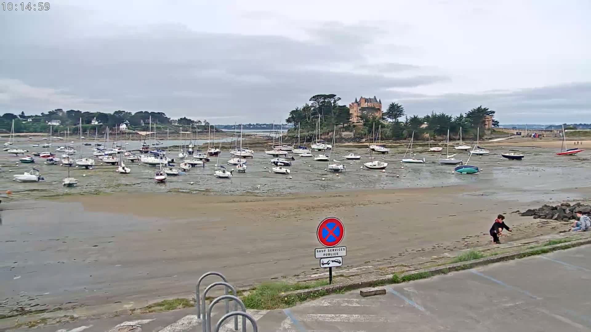 Many sailboats are grounded on a wide, sandy expanse of a bay at low tide under a cloudy sky, near a stone building on a small peninsula.
