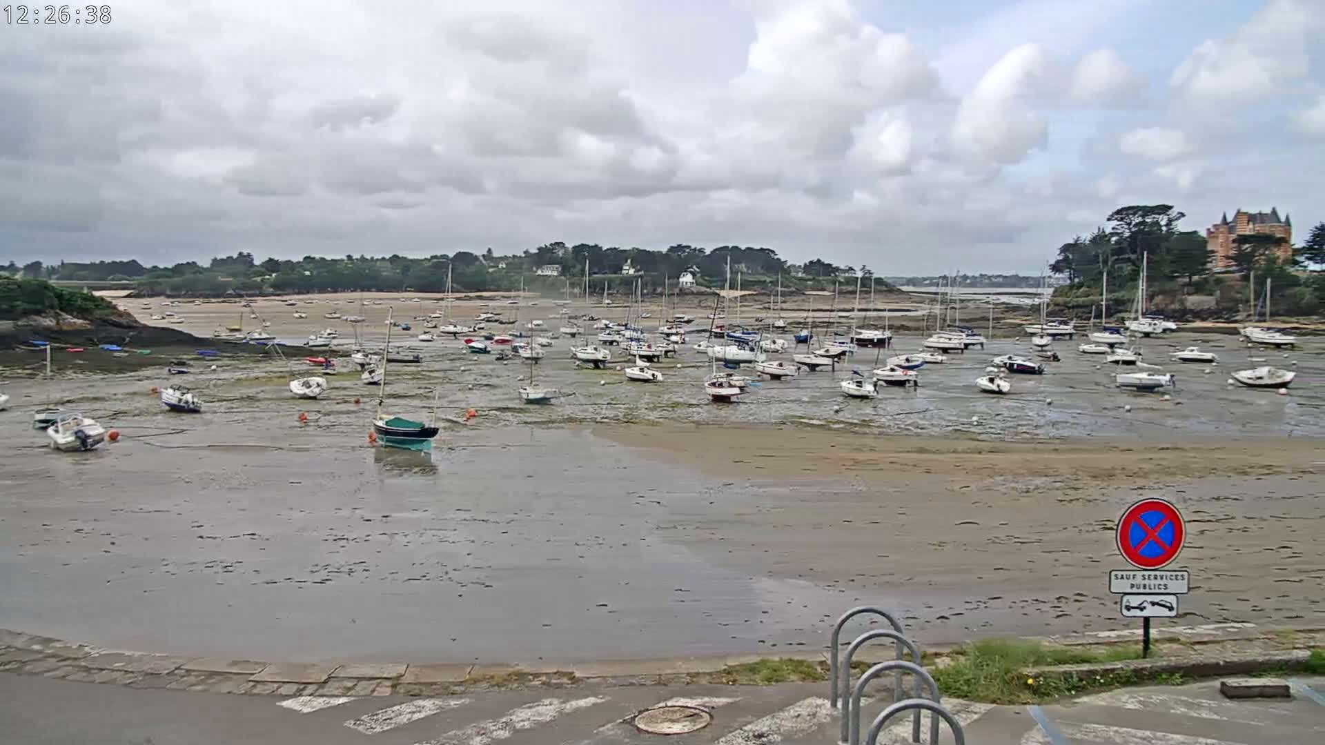 Numerous sailboats are grounded on a wide expanse of exposed mudflats in a harbor under a cloudy sky.