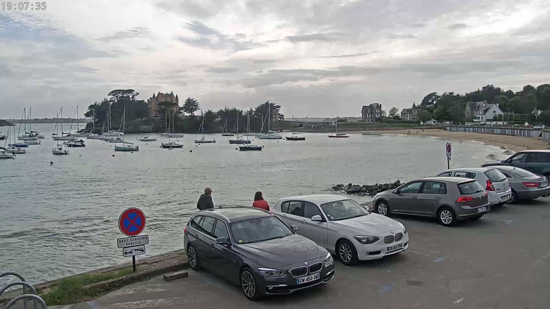 A harbor filled with sailboats is viewed from a parking lot where several cars are parked, under an overcast sky.