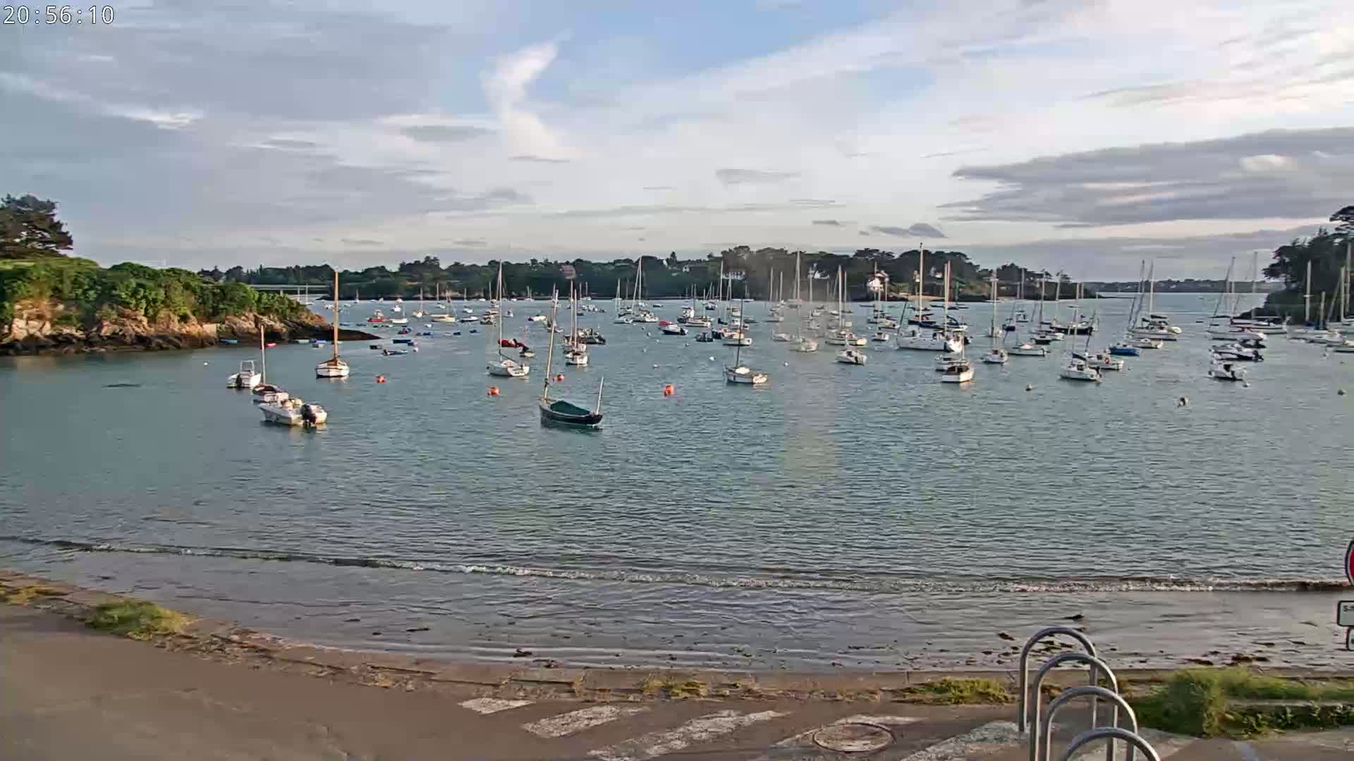 Many sailboats are anchored in a calm bay under a partly cloudy sky.