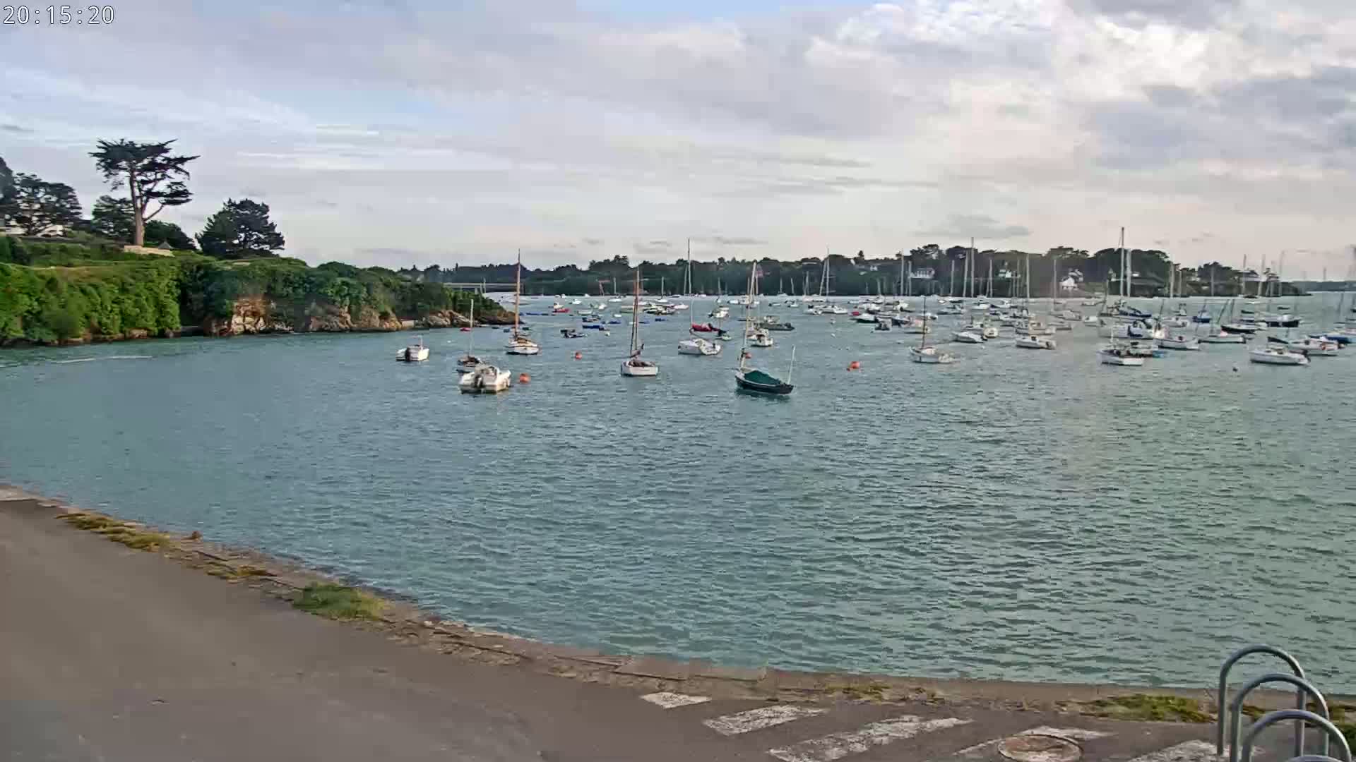 Many sailboats are anchored in a calm bay under a partly cloudy sky.