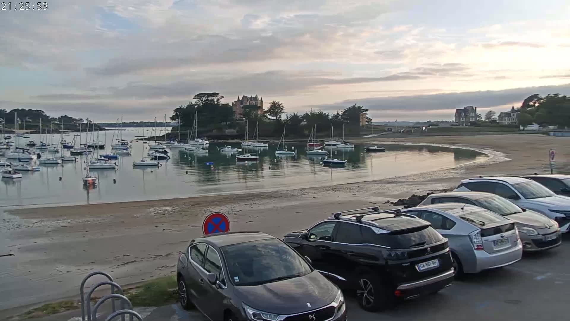 A calm bay with numerous sailboats at anchor, a sandy beach with parked cars, and a chateau on a small hill in the background under a partly cloudy sky.