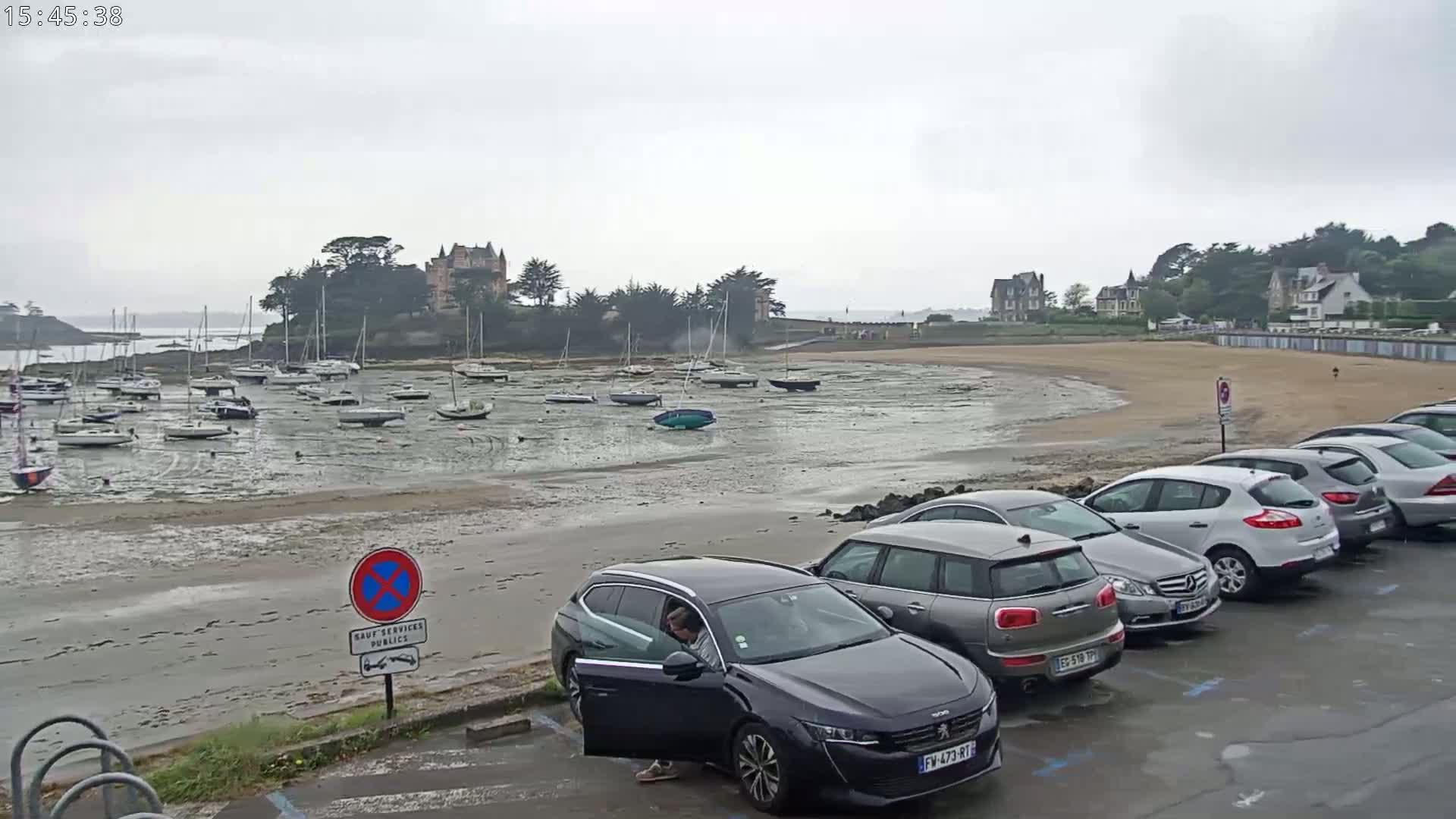 Numerous sailboats are grounded on a tidal flat near a sandy beach and a row of parked cars under an overcast sky.
