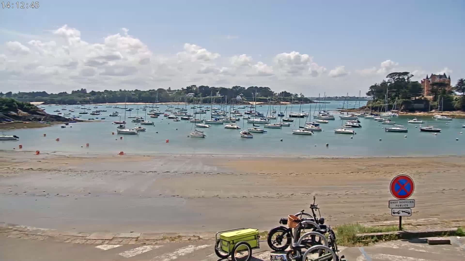 A sandy beach with a calm body of water filled with numerous sailboats, under a partly cloudy sky, and an electric bicycle with a cargo trailer parked in the foreground.