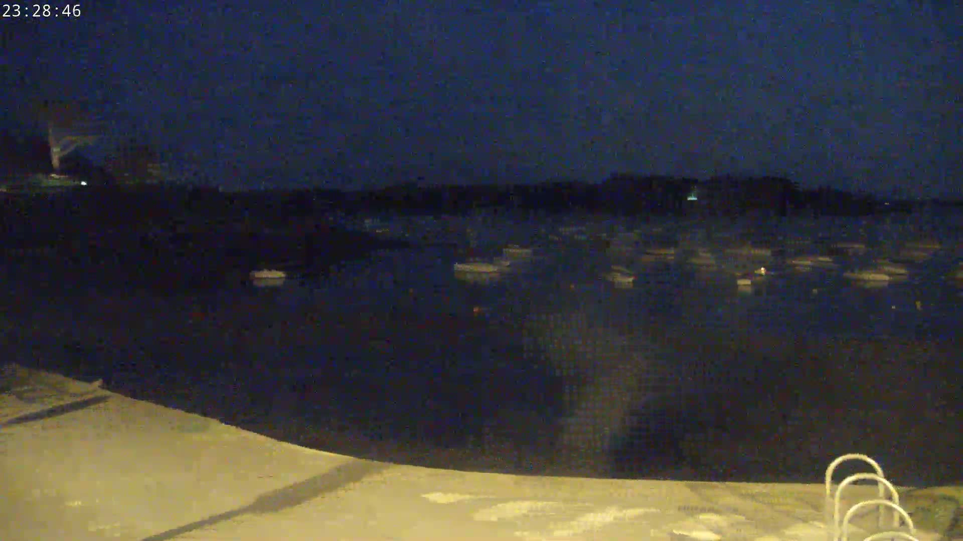 At night, numerous boats are moored in a dark body of water, viewed from a shoreline with steps leading into the water.