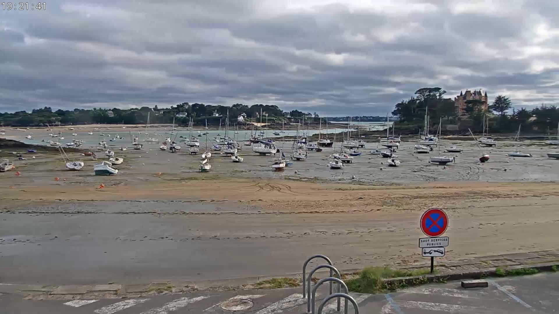 Numerous sailboats are grounded on a sandy expanse of a harbor at low tide under a cloudy sky, with a chateau visible in the background.