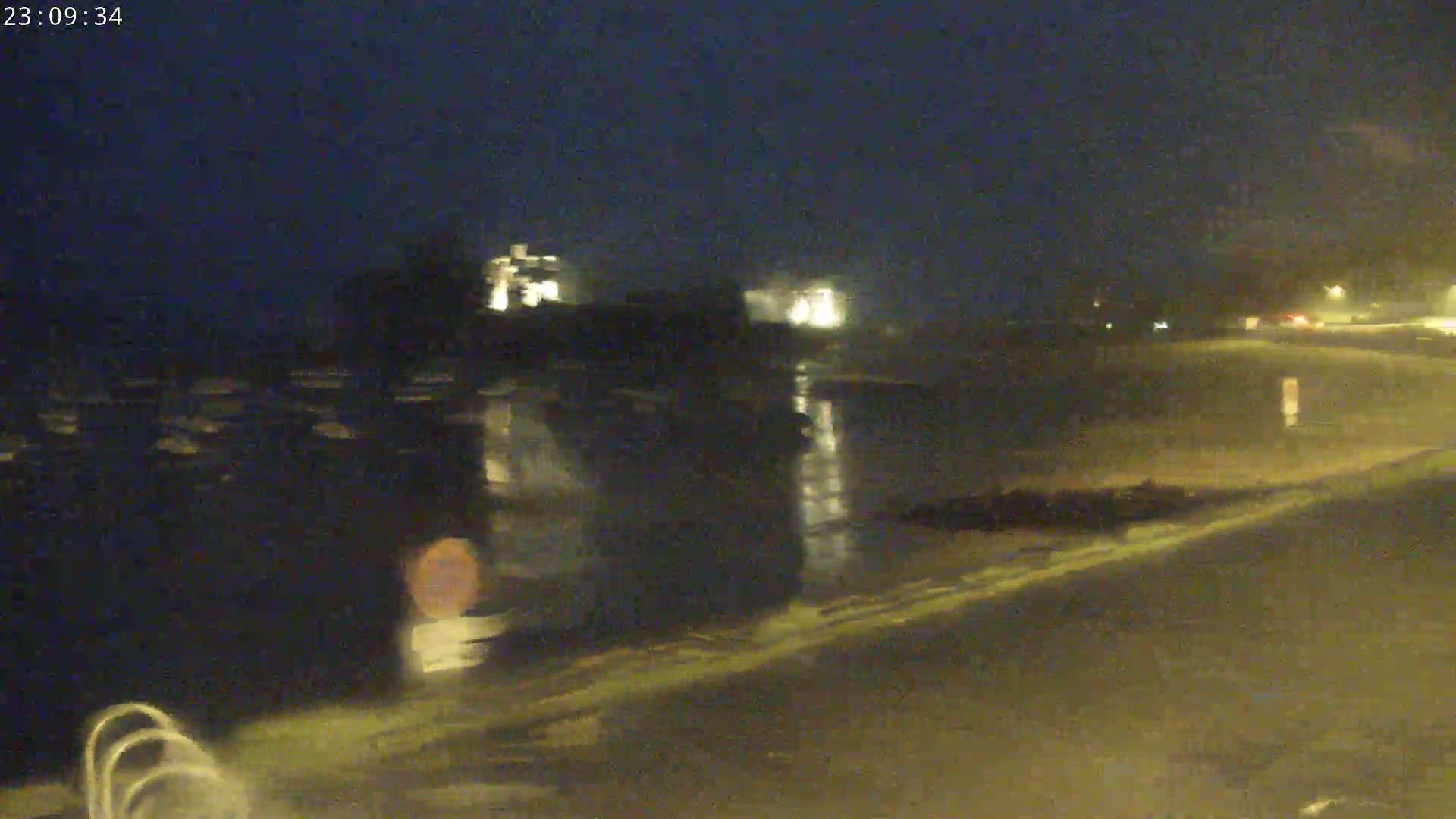A nighttime, wet road scene shows a body of water reflecting lights, with dark buildings and vegetation in the background.