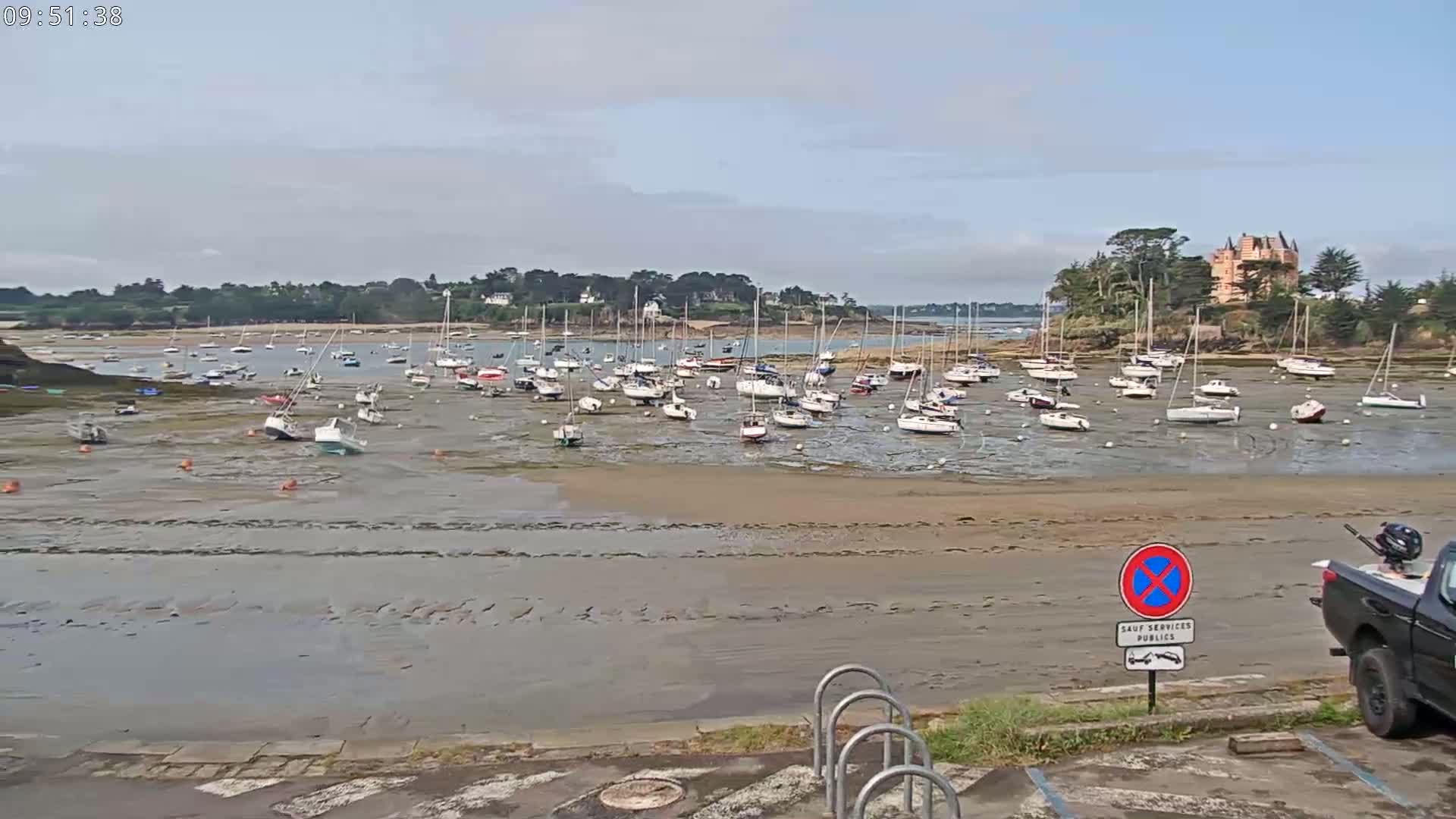Numerous sailboats are grounded on a wide expanse of exposed mudflats in a harbor, under a mostly cloudy sky.
