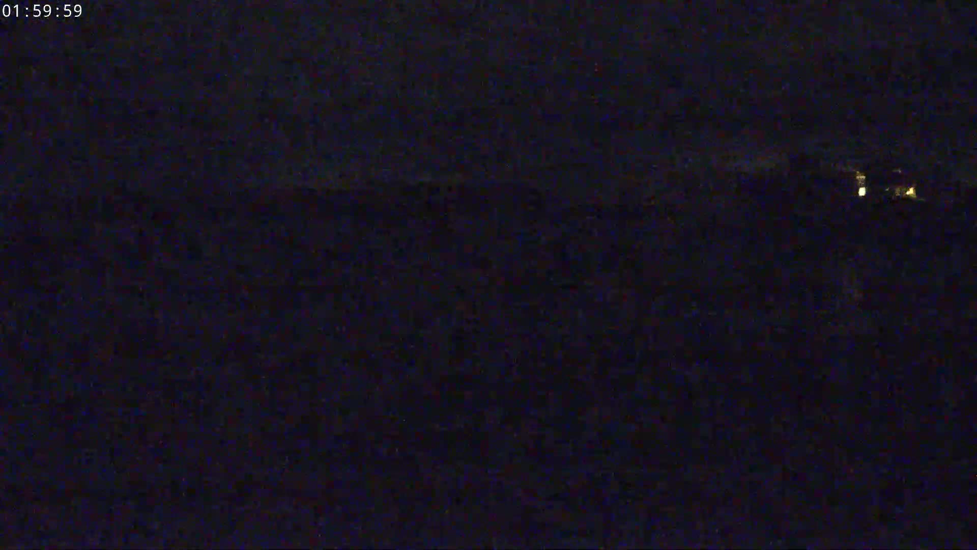 A dark, nighttime scene shows a dimly lit landscape with a few faint lights in the distance under a mostly cloudy sky.