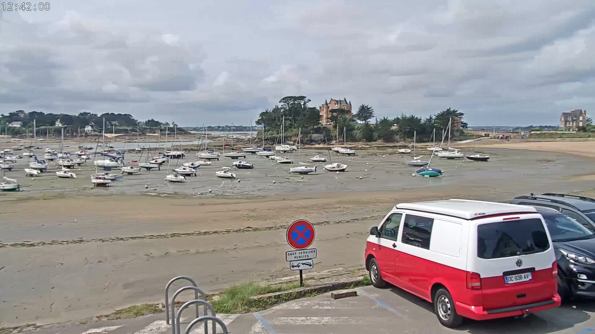 Numerous sailboats are grounded on a wide, exposed sandbar at low tide in front of a chateau on a small island, under a partly cloudy sky.