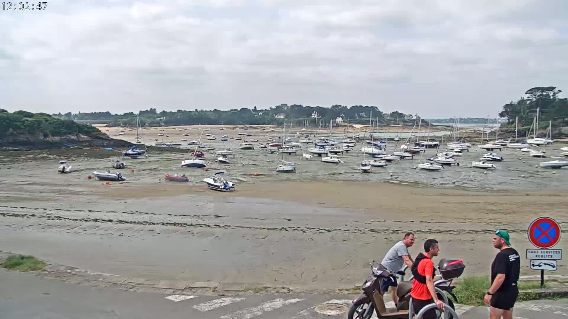 Numerous boats sit on a wide, exposed sandbar at low tide, with three men standing near motor scooters in the foreground under an overcast sky.