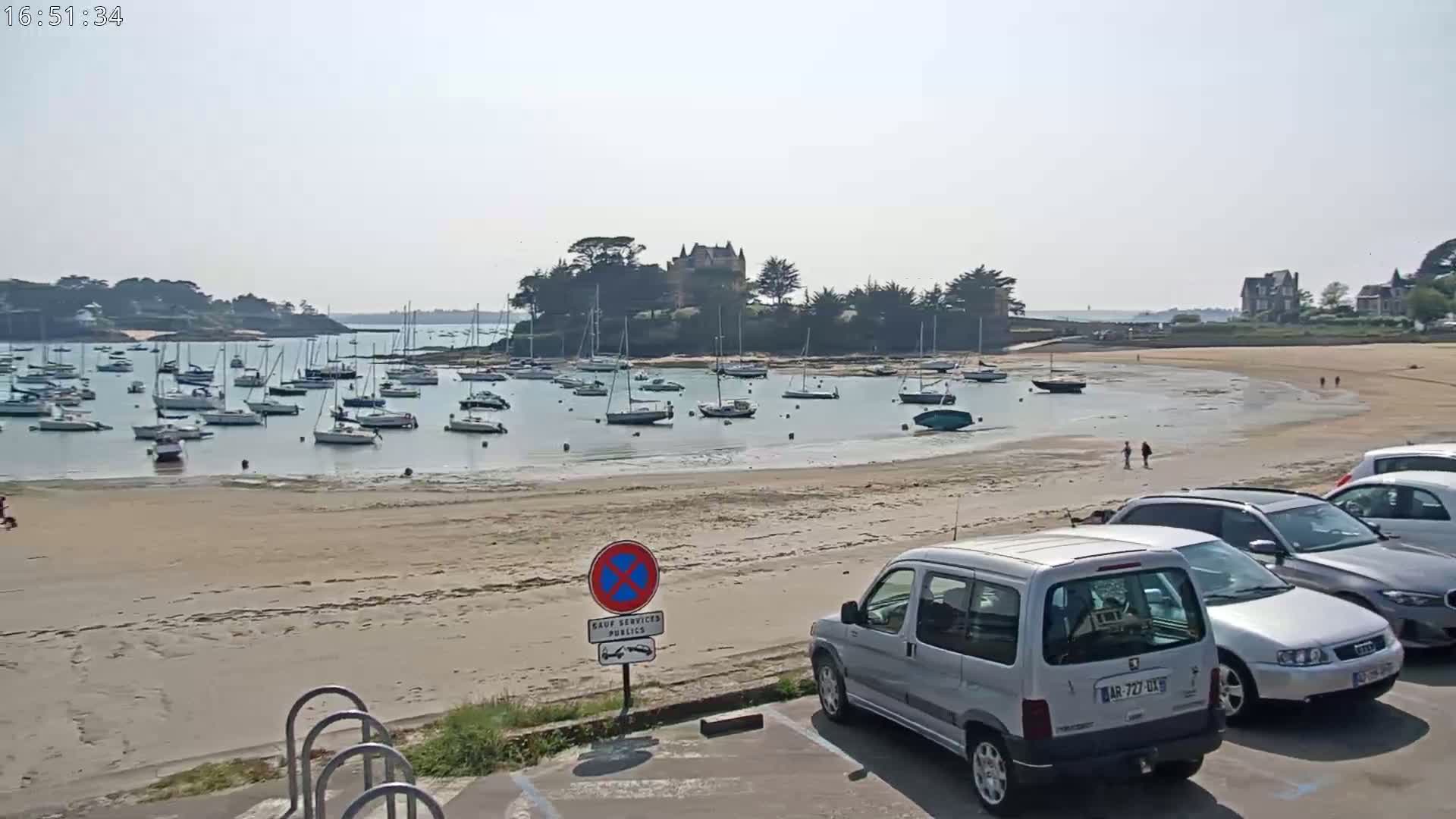 A sandy beach with numerous sailboats in the calm water, a stone building on a small island in the background, and several cars parked nearby in a parking lot under a hazy sky.