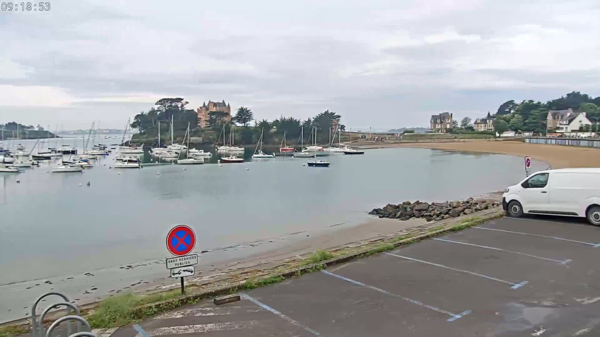 A calm harbor filled with sailboats sits under an overcast sky, next to a sandy beach and a parking lot with a single white van.