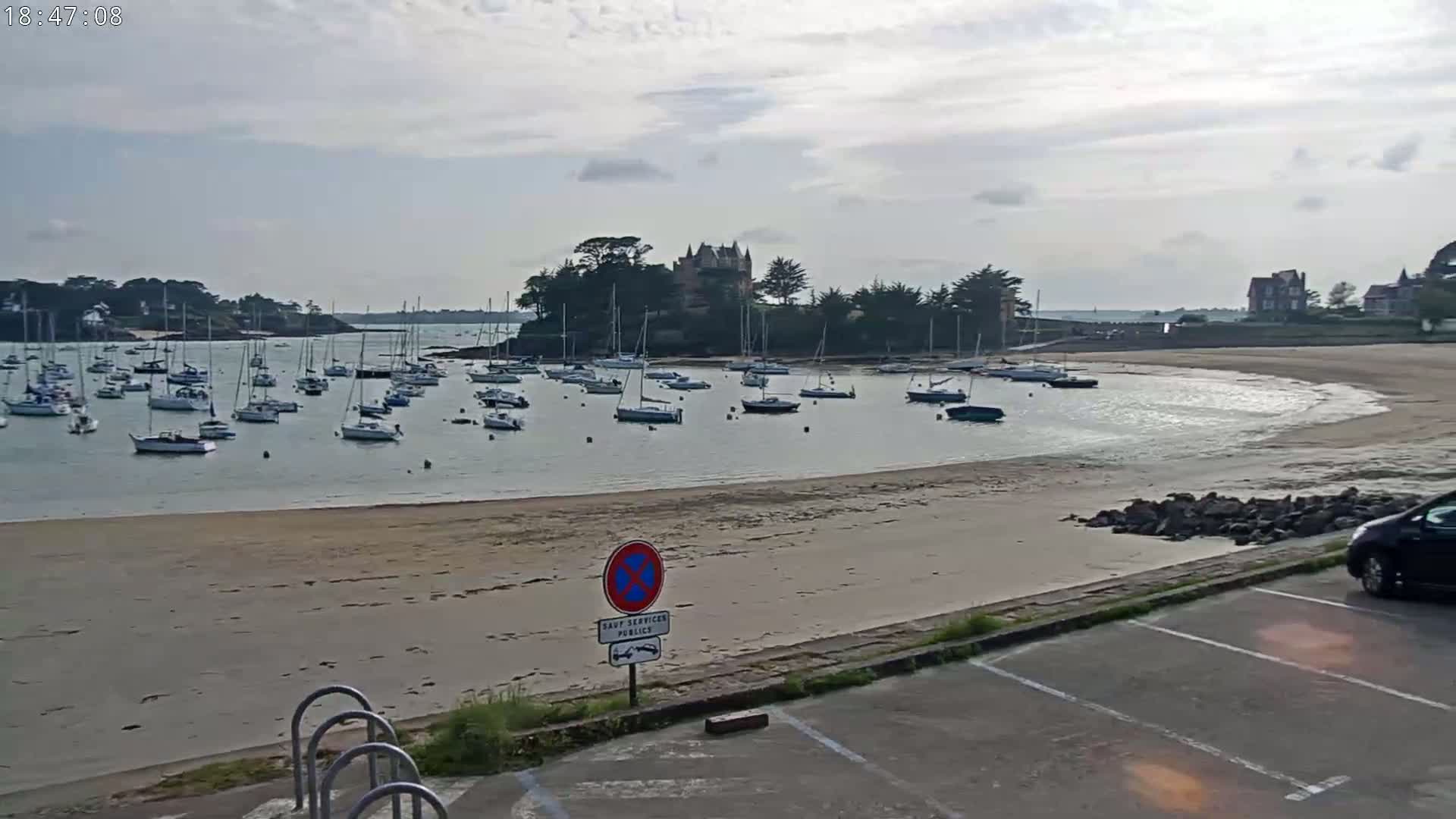A calm, overcast day reveals a sandy beach, a harbor filled with sailboats, and a large building perched on a small, tree-covered island.