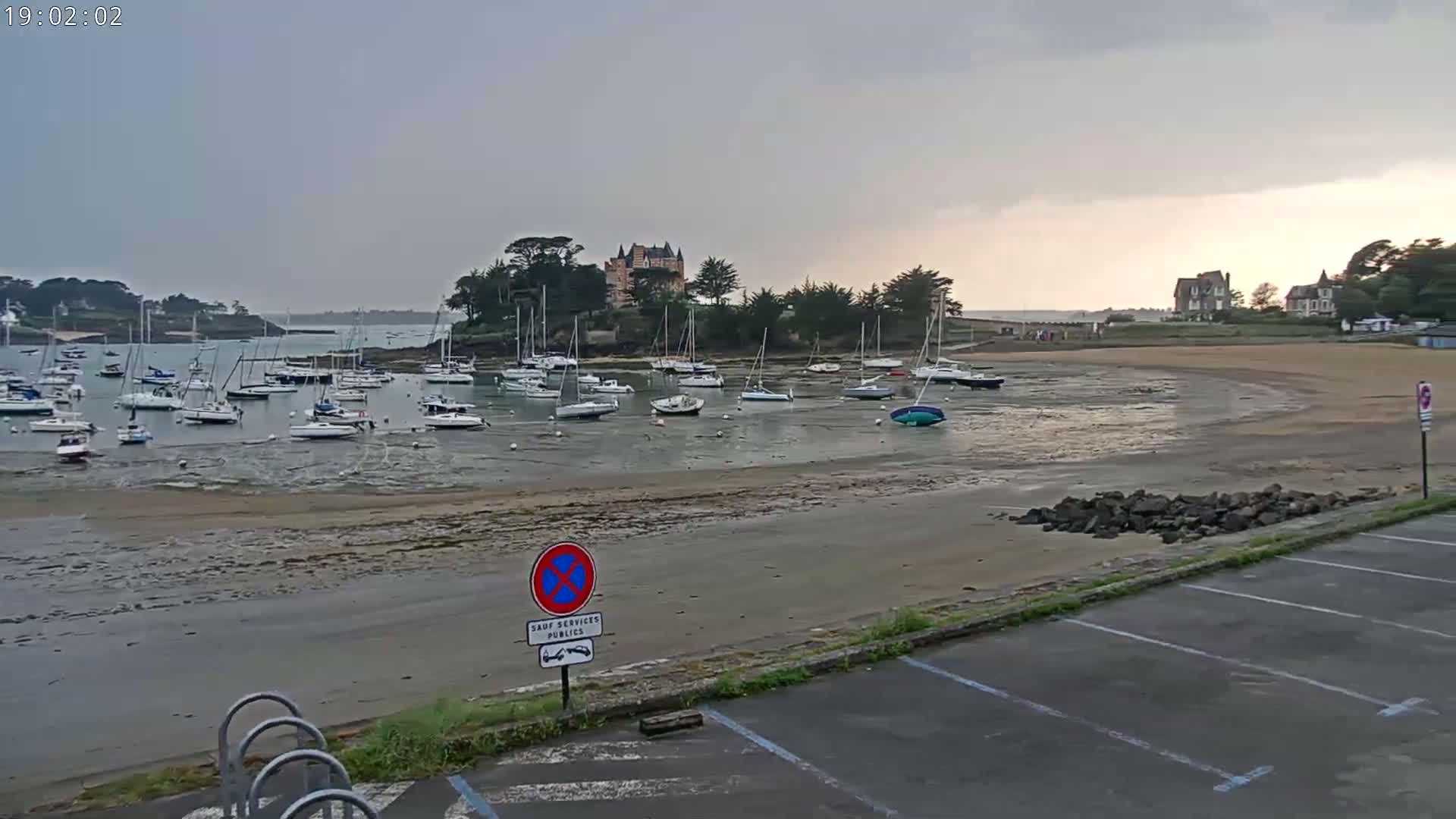 A low-tide harbor filled with sailboats sits in front of a sandy beach and a chateau, under an overcast sky.