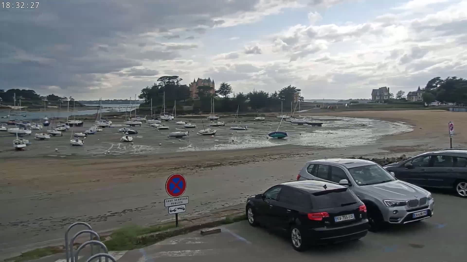 Many sailboats are grounded on a sandy shore at low tide under a cloudy sky, with several cars parked nearby.