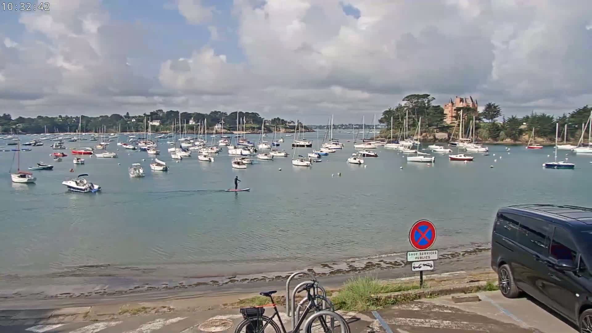 A calm bay filled with numerous sailboats and motorboats is viewed from a shoreline where a person paddleboards and bicycles are parked near a dark-colored van under a partly cloudy sky.