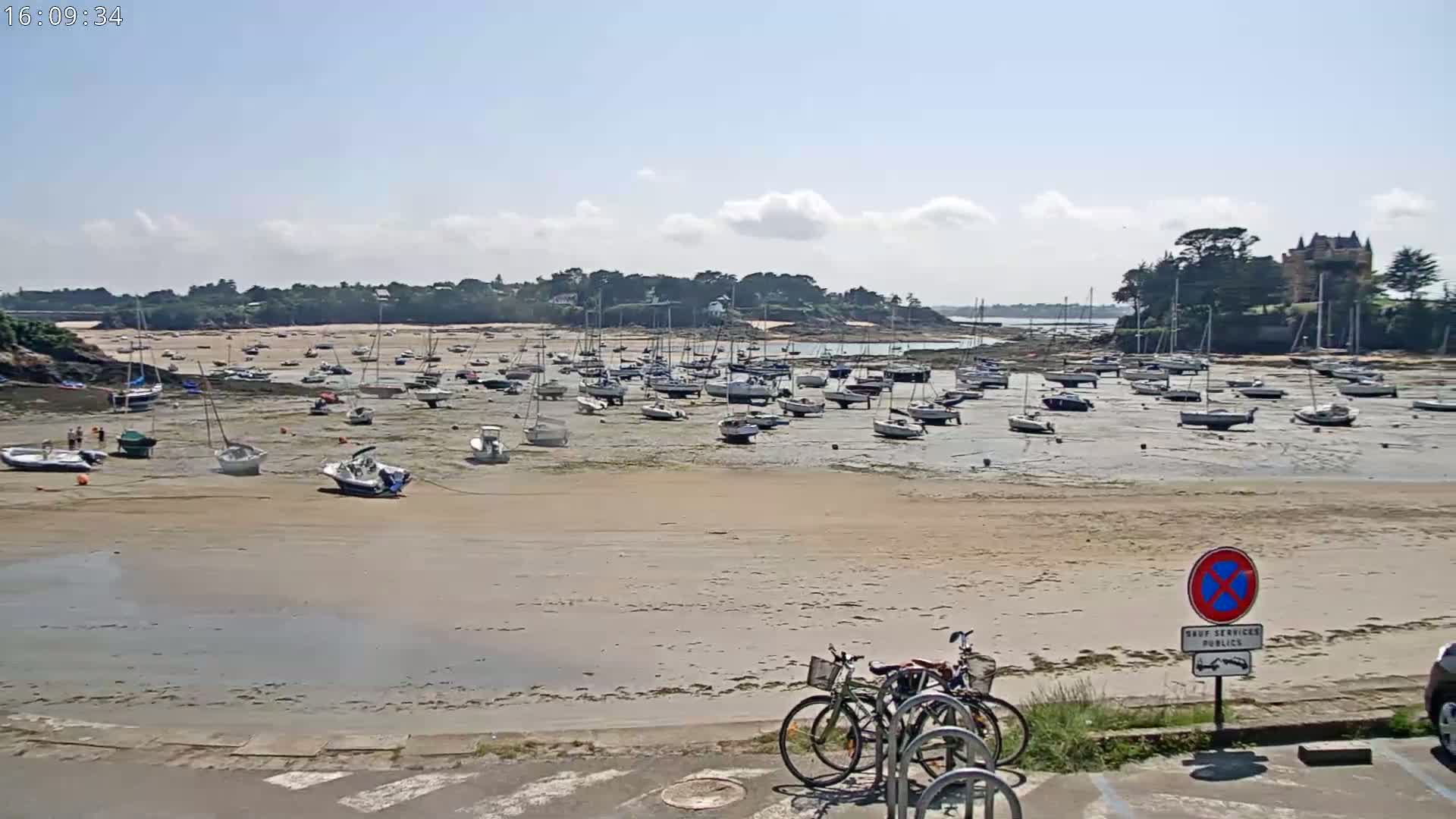 Numerous sailboats are grounded on a wide, sandy beach at low tide under a mostly sunny sky.