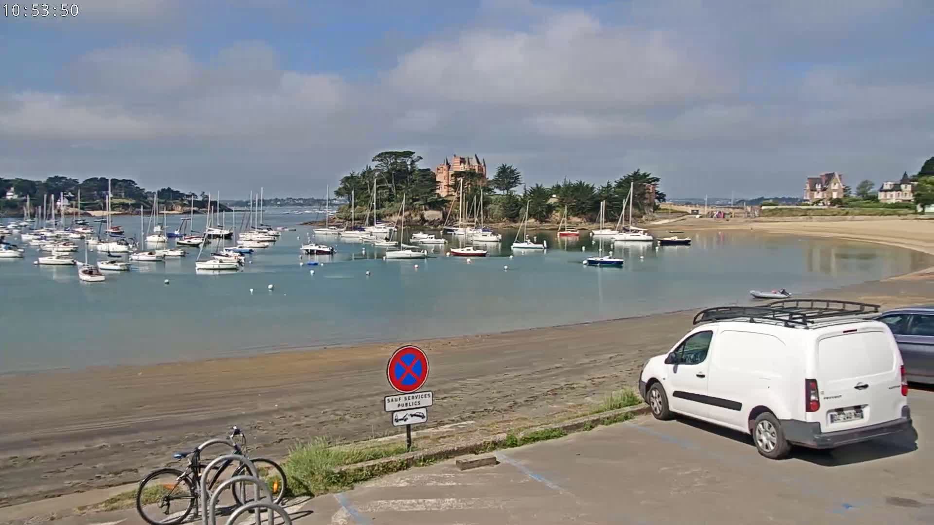 A calm bay with numerous sailboats is viewed from a sandy shore on a partly sunny day, with a white van parked in a nearby lot.