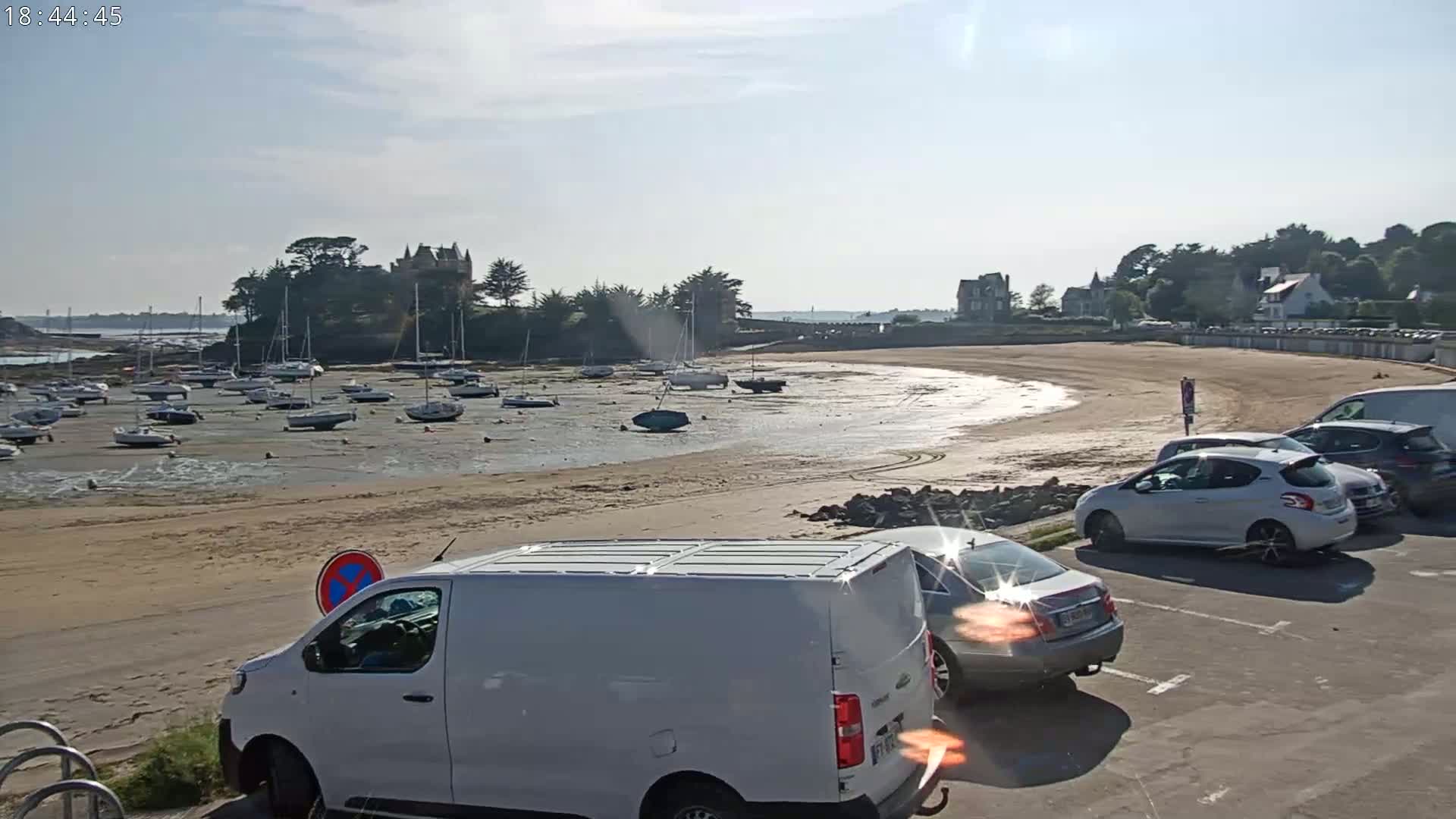 A sandy beach with numerous sailboats grounded at low tide, cars parked in a lot, and a stone structure on a small island in the background under a partly sunny sky.