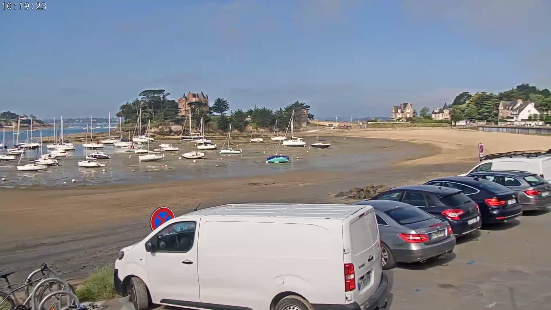 A sandy beach with numerous sailboats grounded at low tide, several cars parked nearby, and a chateau visible in the background under a clear blue sky.