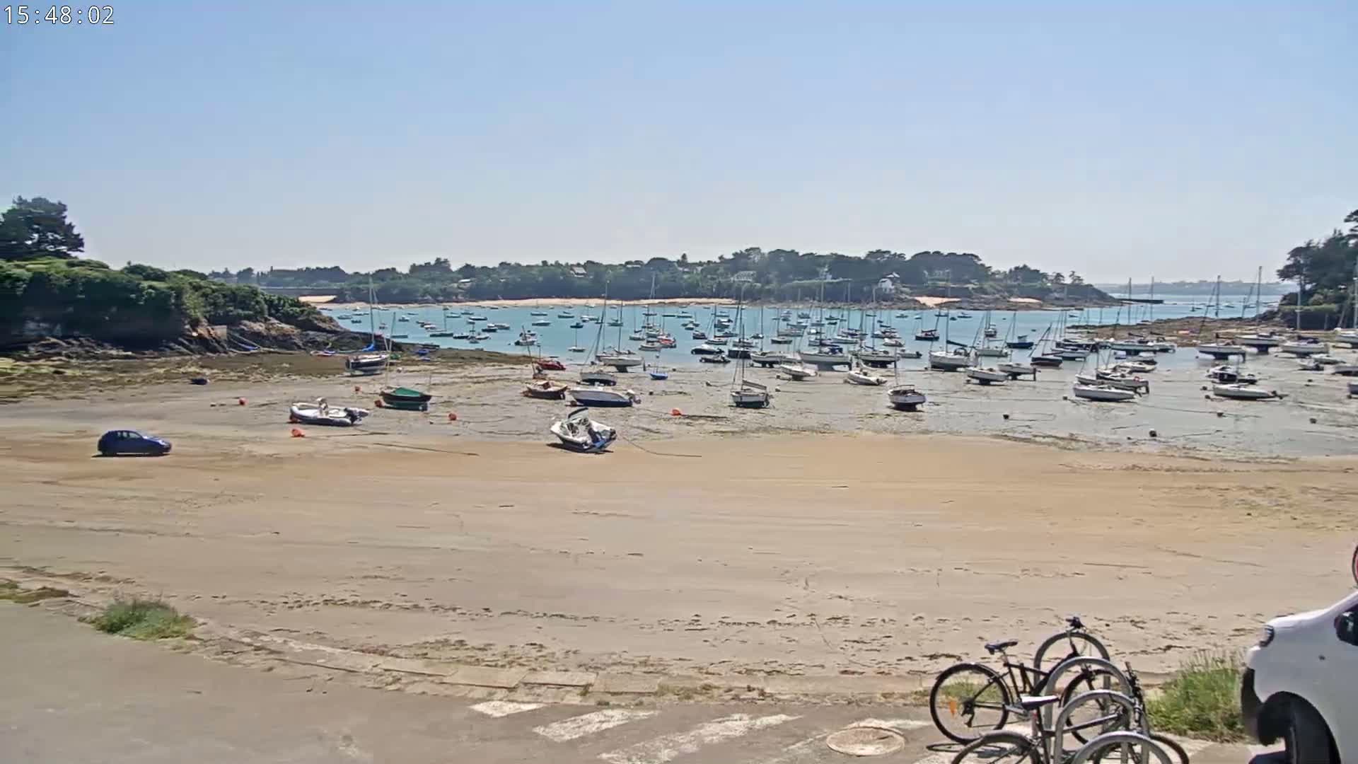 A sandy beach with numerous boats grounded at low tide, a small car, and bicycles parked near a roadway, under sunny skies.