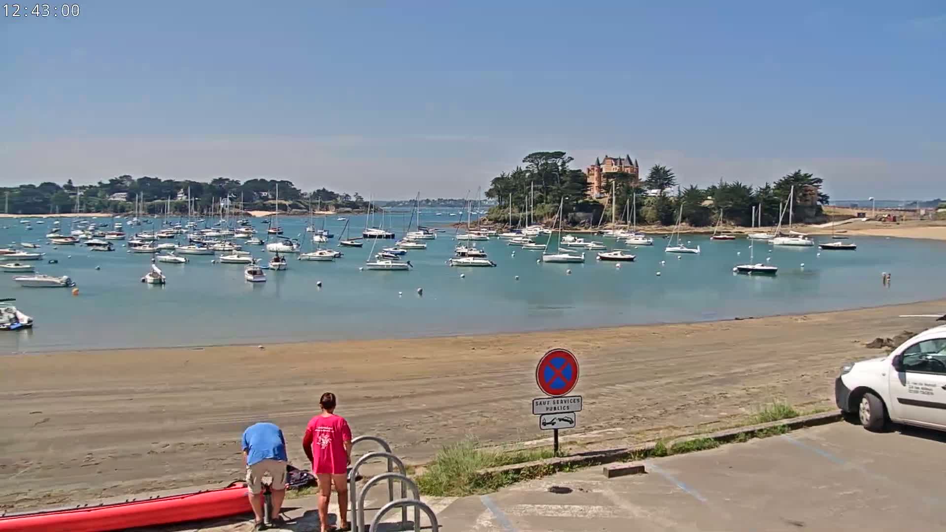 A sandy beach overlooks a calm bay filled with sailboats, under a clear blue sky, with two people near a red kayak and a white van parked nearby.
