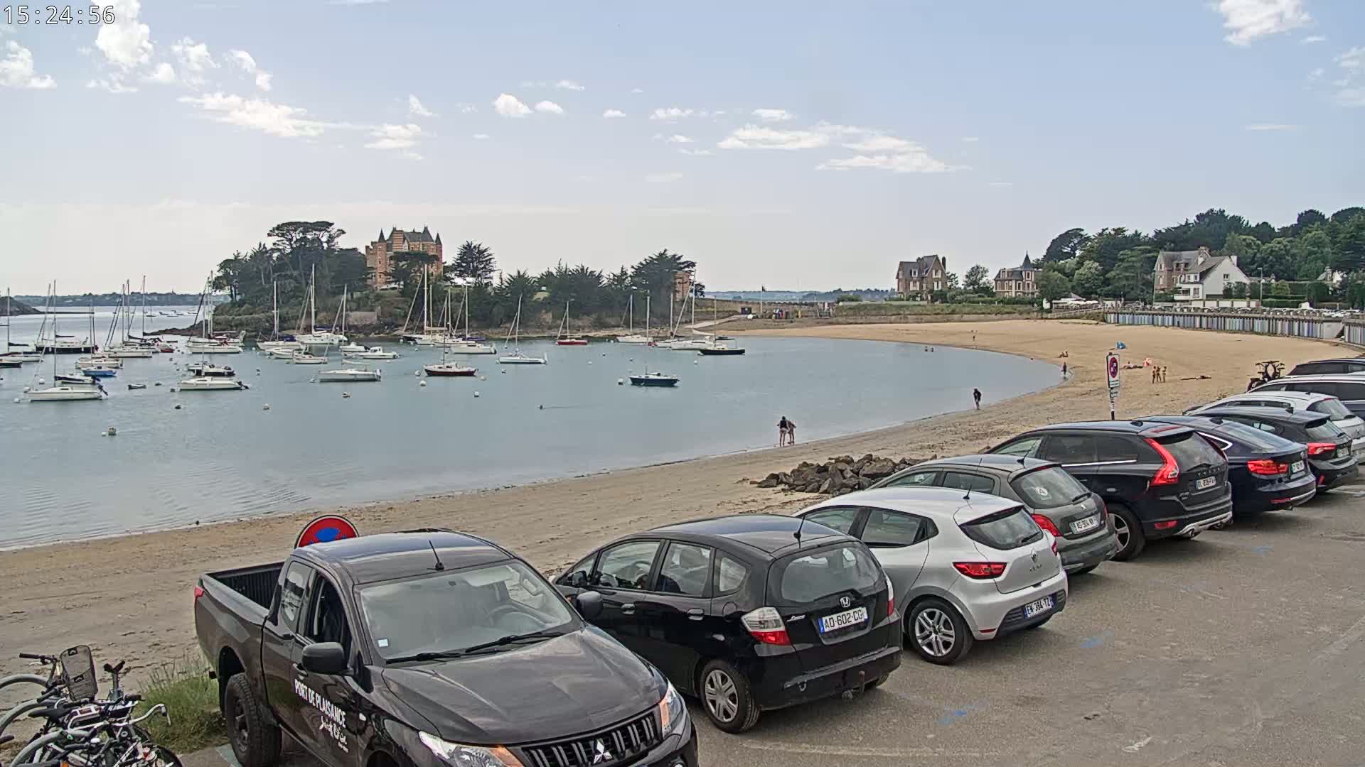 A sandy beach with parked cars and a calm harbor filled with sailboats, under a mostly sunny sky.