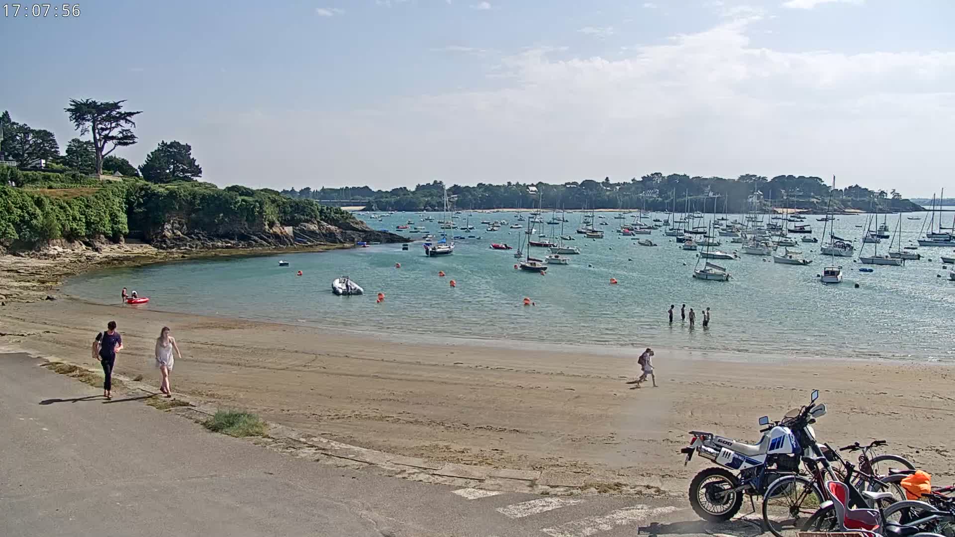 A sandy beach with a few people walking and swimming, many sailboats anchored in calm, clear water under a sunny sky.