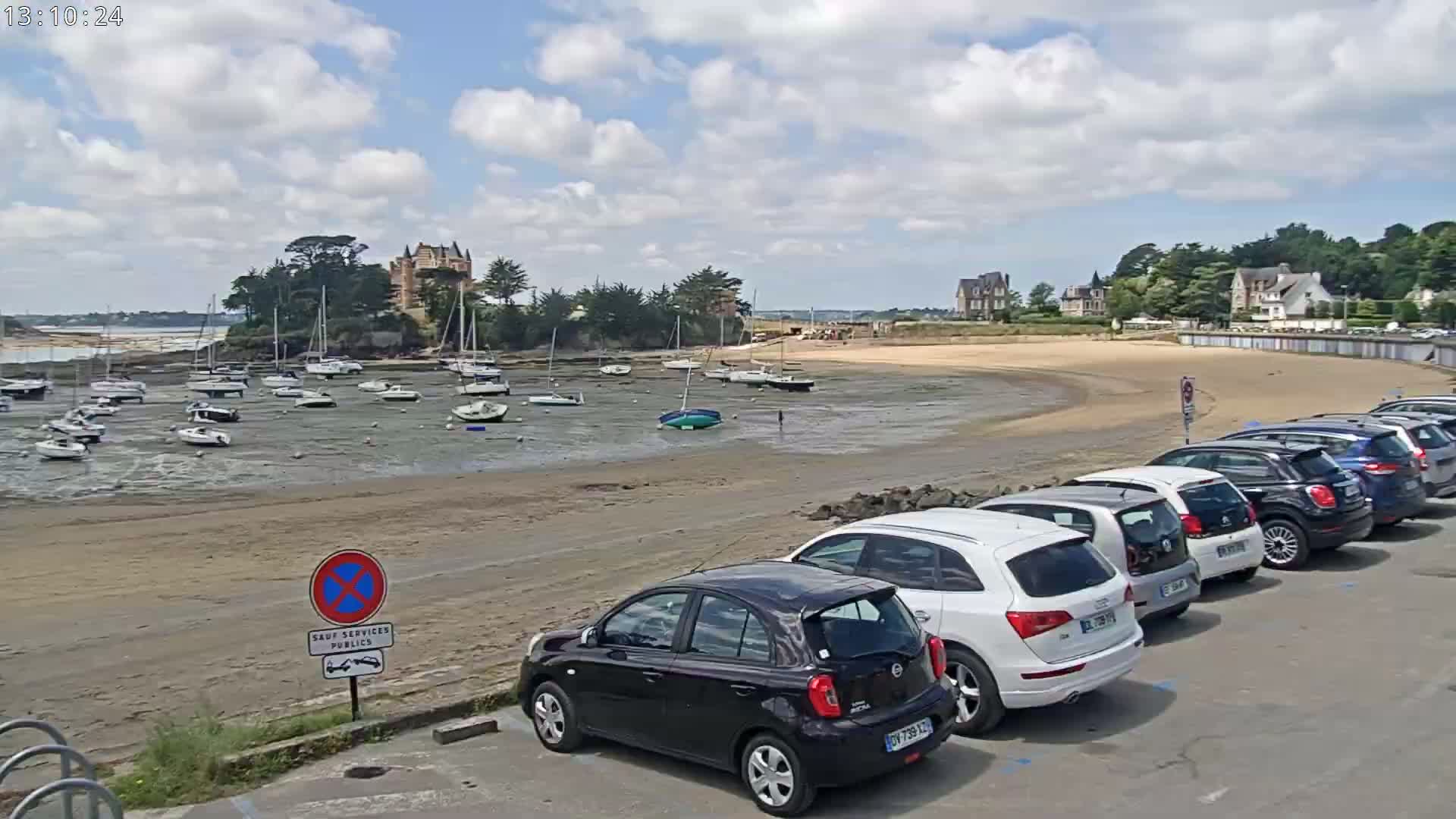 A sandy beach with numerous sailboats at low tide is bordered by a row of parked cars,  a stone building, and lush green trees under a partly cloudy sky.