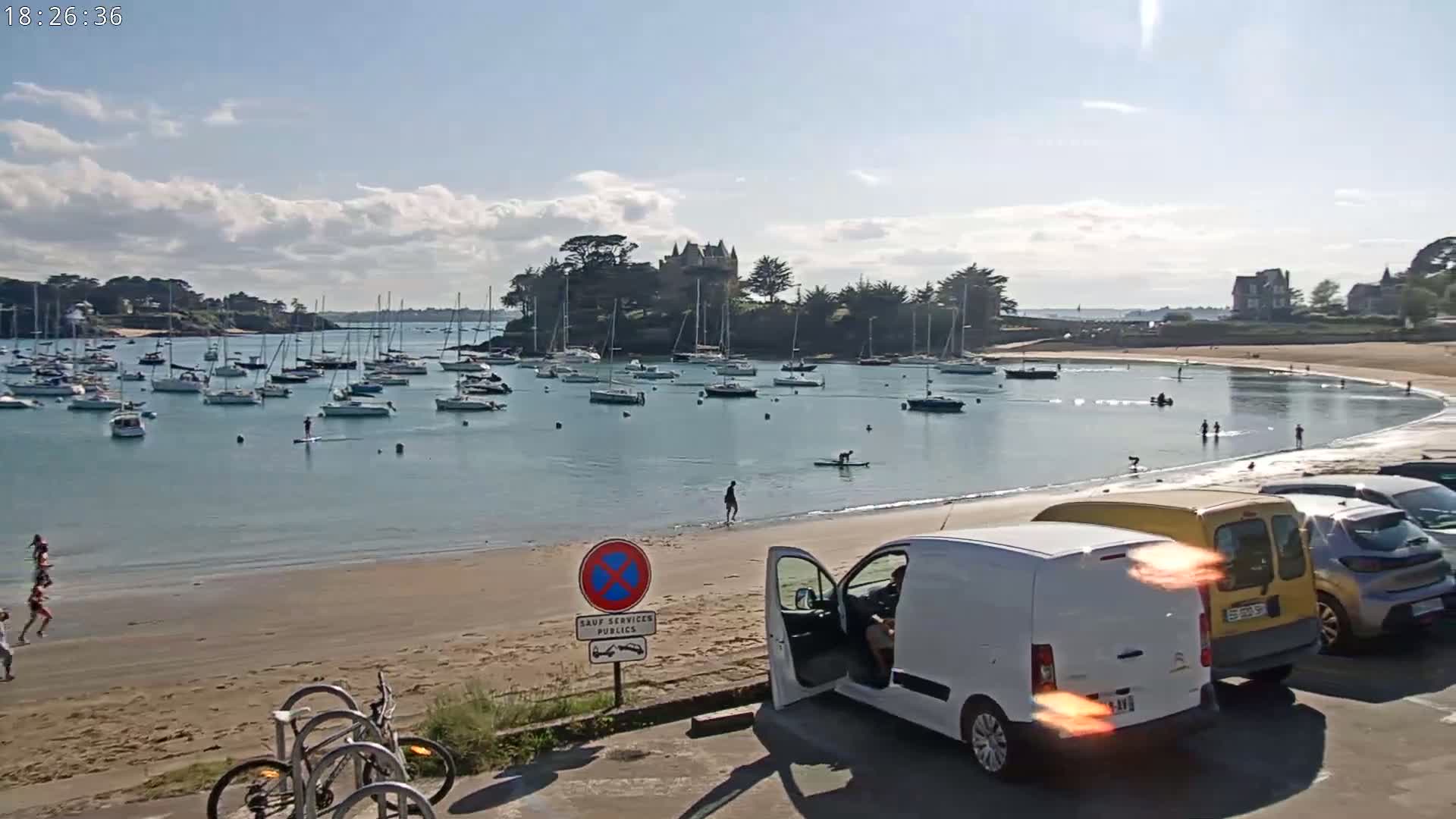 A sandy beach with calm, clear water, numerous sailboats in a harbor, a few people swimming and walking along the shore, and cars parked nearby under a partly cloudy sky.