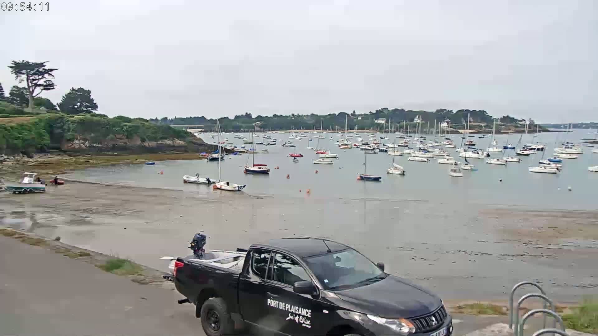 A calm, overcast day reveals a harbor filled with numerous sailboats, a sandy shore, and a dark-colored pickup truck parked near a paved road.