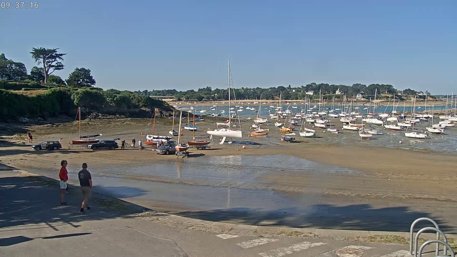 A sunny day reveals a sandy harbor with numerous sailboats at low tide, several cars parked near the shore, and a few people standing nearby.