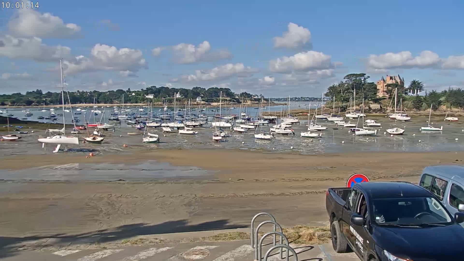 Many sailboats are docked in shallow water on a sunny day with partly cloudy skies, near a shoreline with a building and vehicles parked nearby.