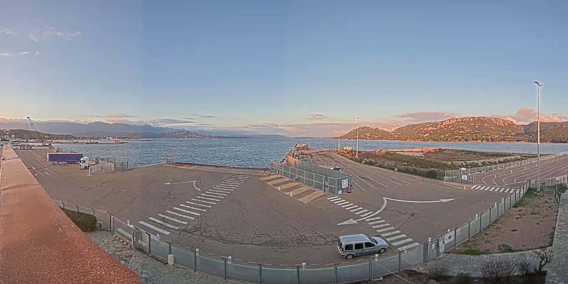 A mostly empty ferry terminal with a single vehicle parked in the lot overlooks a calm body of water under a clear, sunset sky with mountains in the distance.