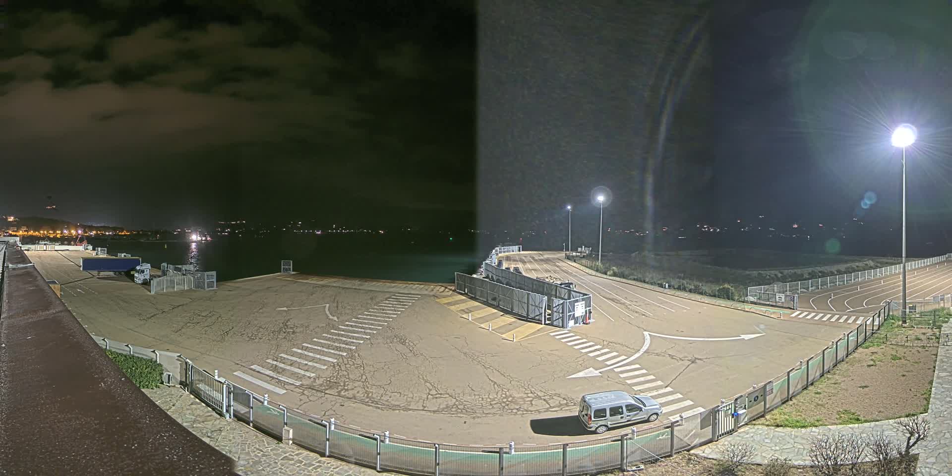 A nighttime view of an empty port area with a single vehicle parked, under a cloudy sky, next to a track and field.