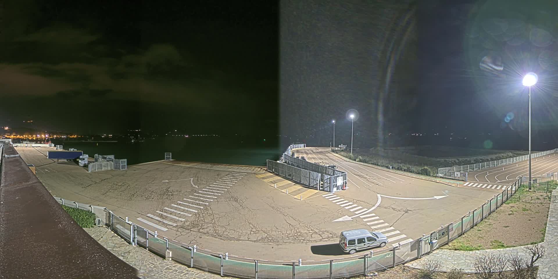 A nighttime view of a mostly empty port area with a van parked near a fenced-in track, under a partly cloudy sky.