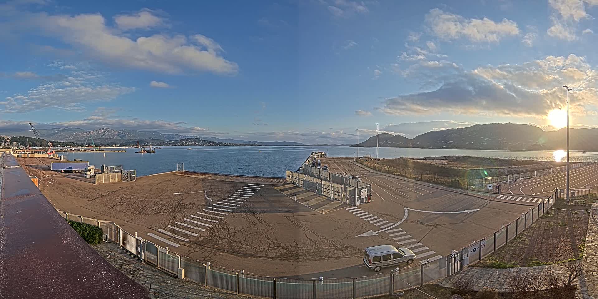 A mostly empty port area with a vehicle and a partially cloudy sky over calm water and distant mountains.