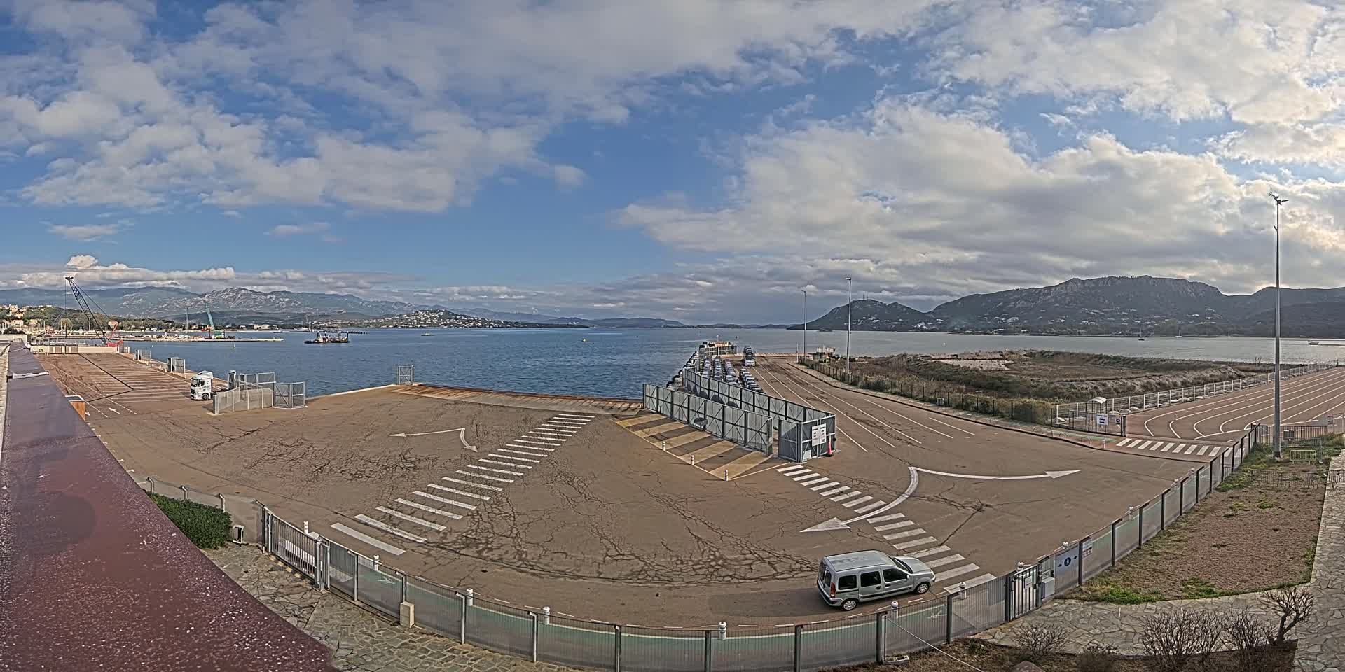 A mostly empty ferry terminal with a few vehicles, under a partly cloudy sky, overlooks a calm body of water and distant mountains.