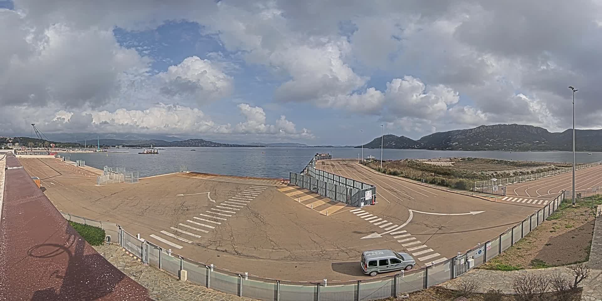 An empty port area with a single vehicle, under a partly cloudy sky, overlooks calm water and distant hills.