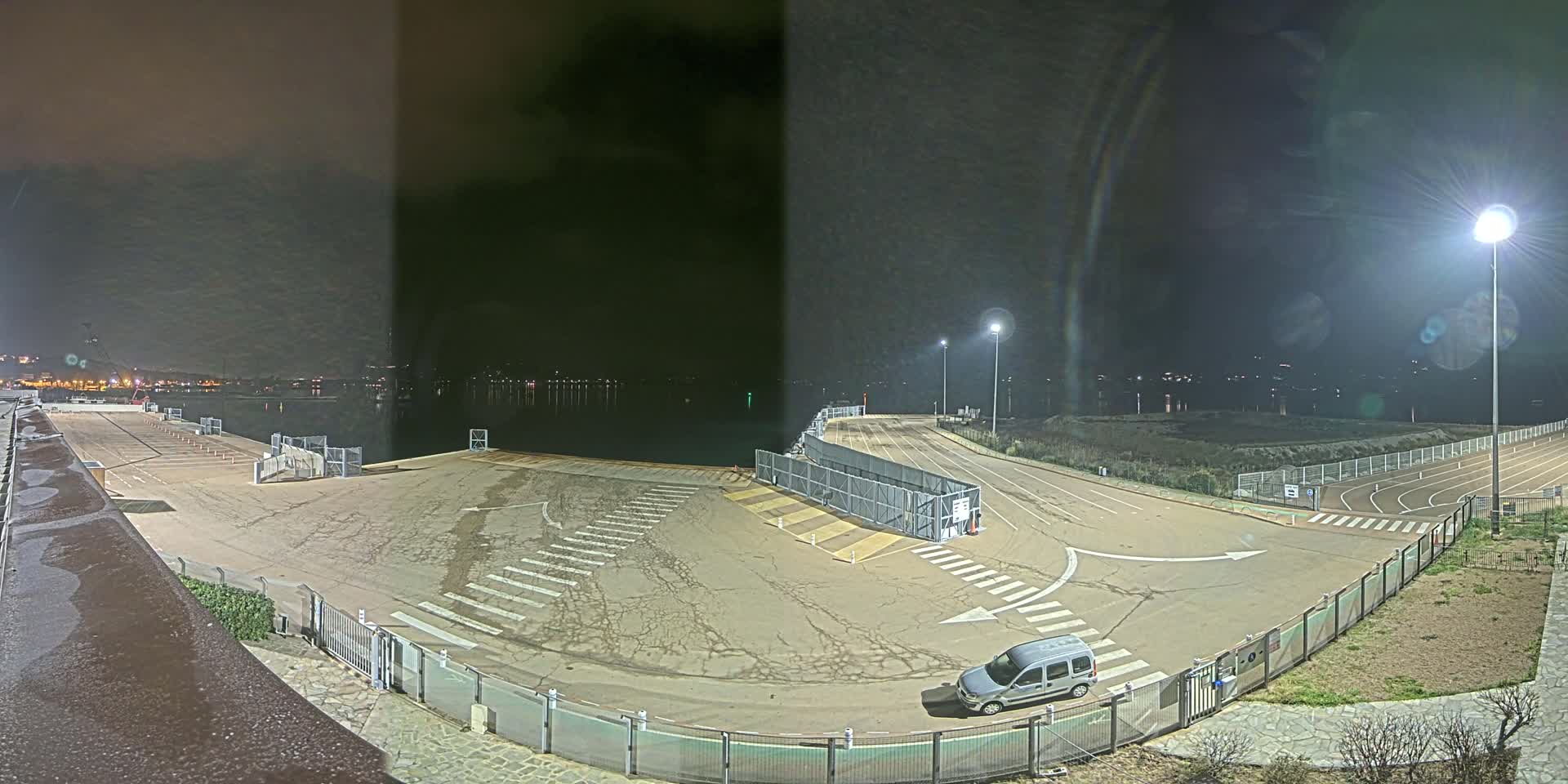 A nighttime view of an empty parking lot and roadway near a body of water, illuminated by artificial lights.
