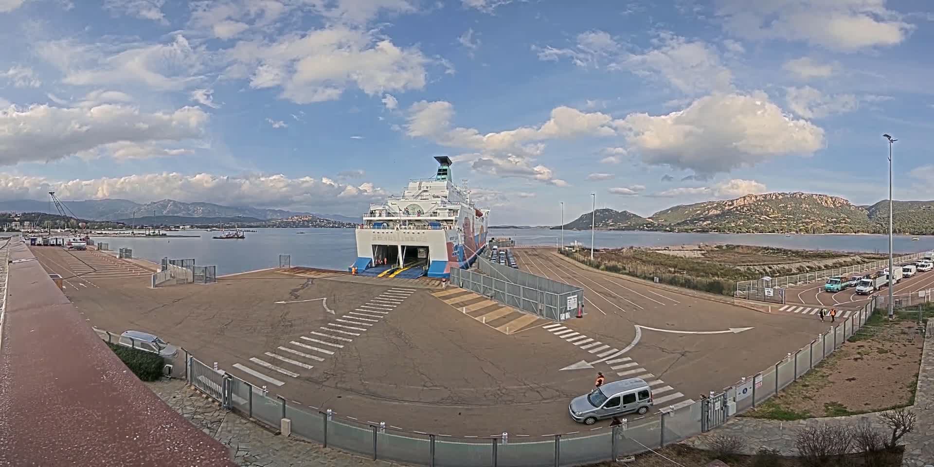 A large ferry is docked at a port under a partly cloudy sky, with several vehicles parked nearby and mountains visible in the distance.