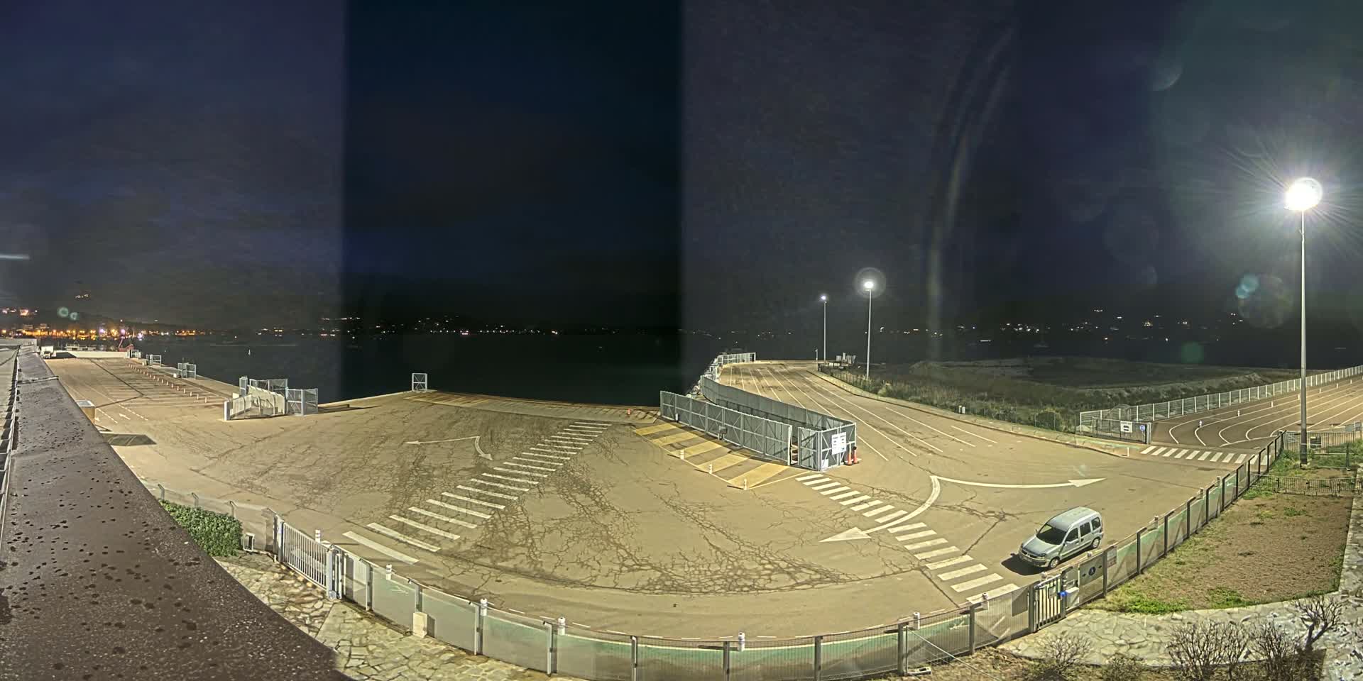 An empty, dark asphalt area with marked lanes and a single van, next to a body of water under a night sky.