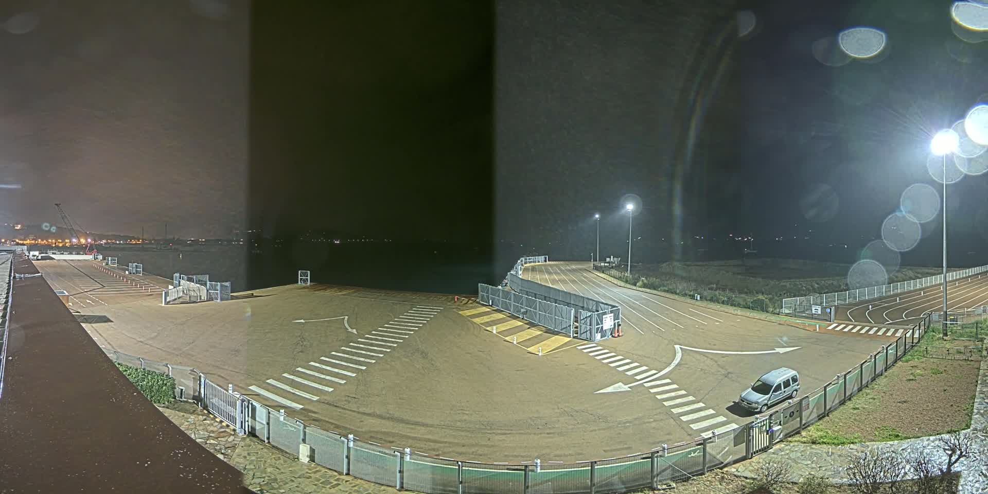 A nighttime view of an empty port area with a single van parked near a fenced-in section, under slightly overcast skies and rain.