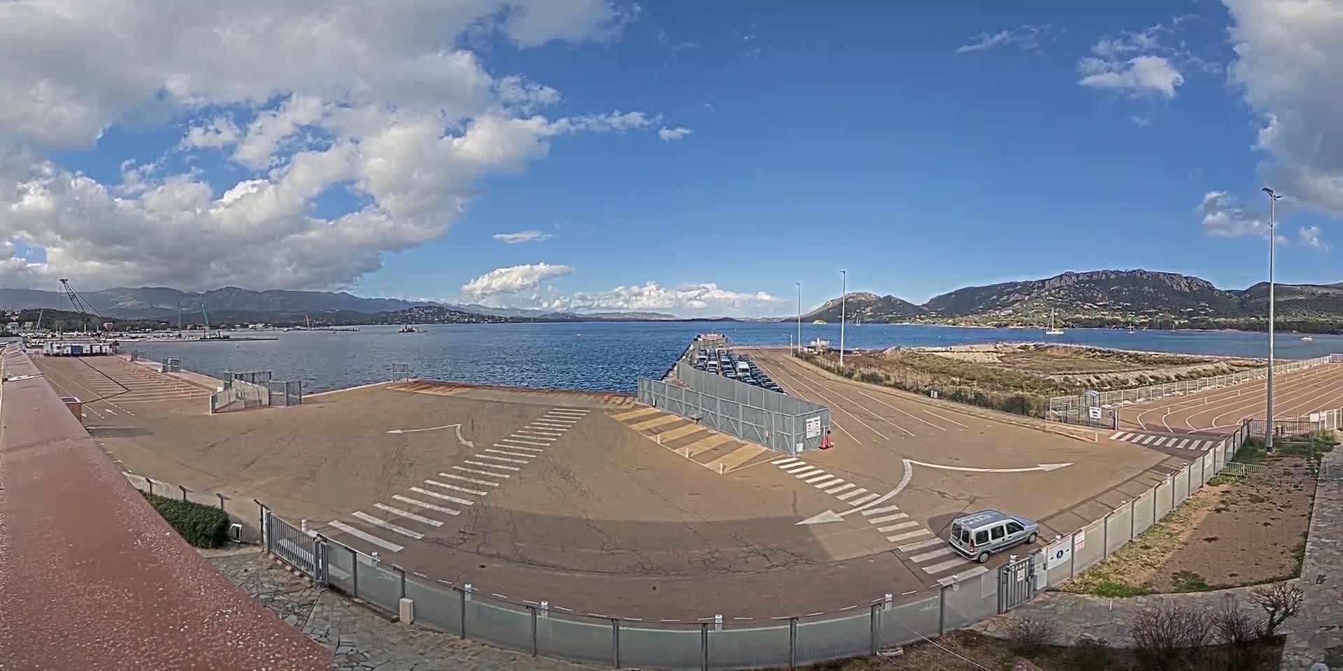 A mostly sunny day shows a ferry terminal with a mostly empty parking lot, a line of cars waiting to board, and calm blue water with mountains in the distance.