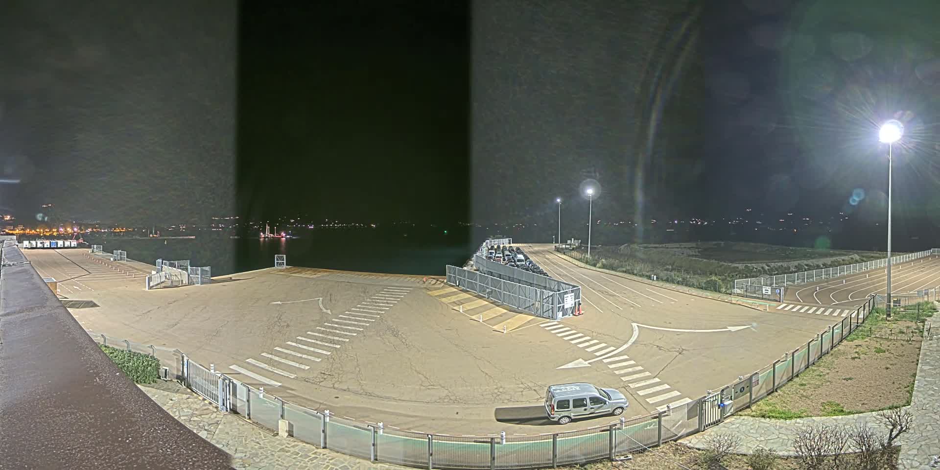 An empty parking lot at night next to a body of water, illuminated by several bright lights, with a single vehicle parked near a gated area.